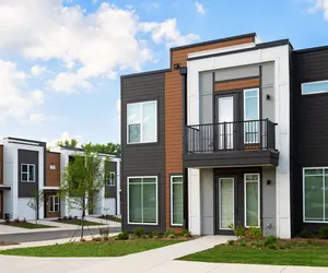 A modern apartment complex featuring sleek architectural design with a combination of dark and light colored facades, green landscaping, and a small pathway in front.