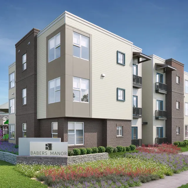 A modern apartment building with a mix of light and dark exterior colors, featuring large windows. Flower beds with colorful plants are in the foreground, and a sign with 'Barber's Manor' is visible near the entrance. There are trees and additional buildings in the background under a clear blue sky.
