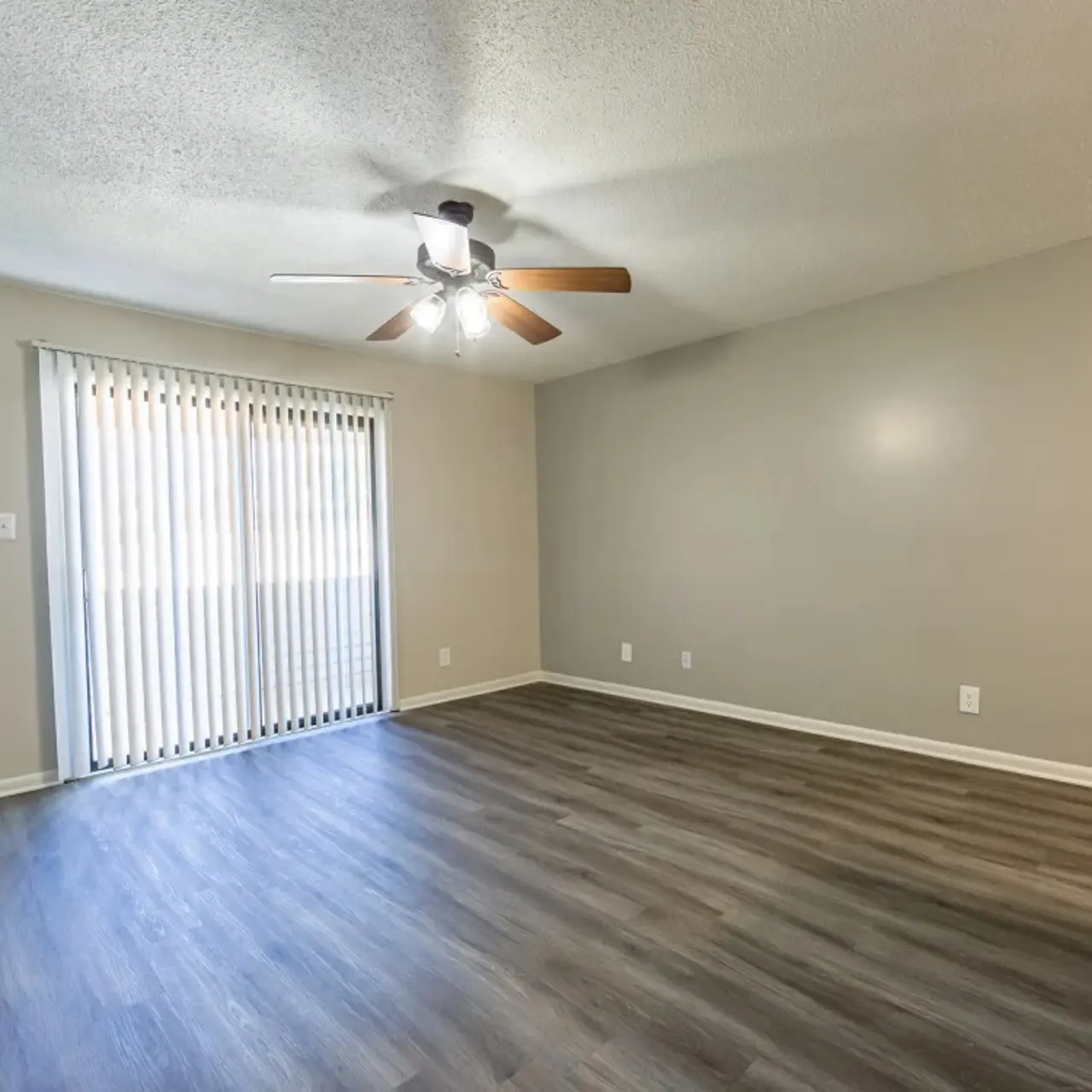 A spacious living room with a ceiling fan, light wood-like flooring, and a large window with vertical blinds.