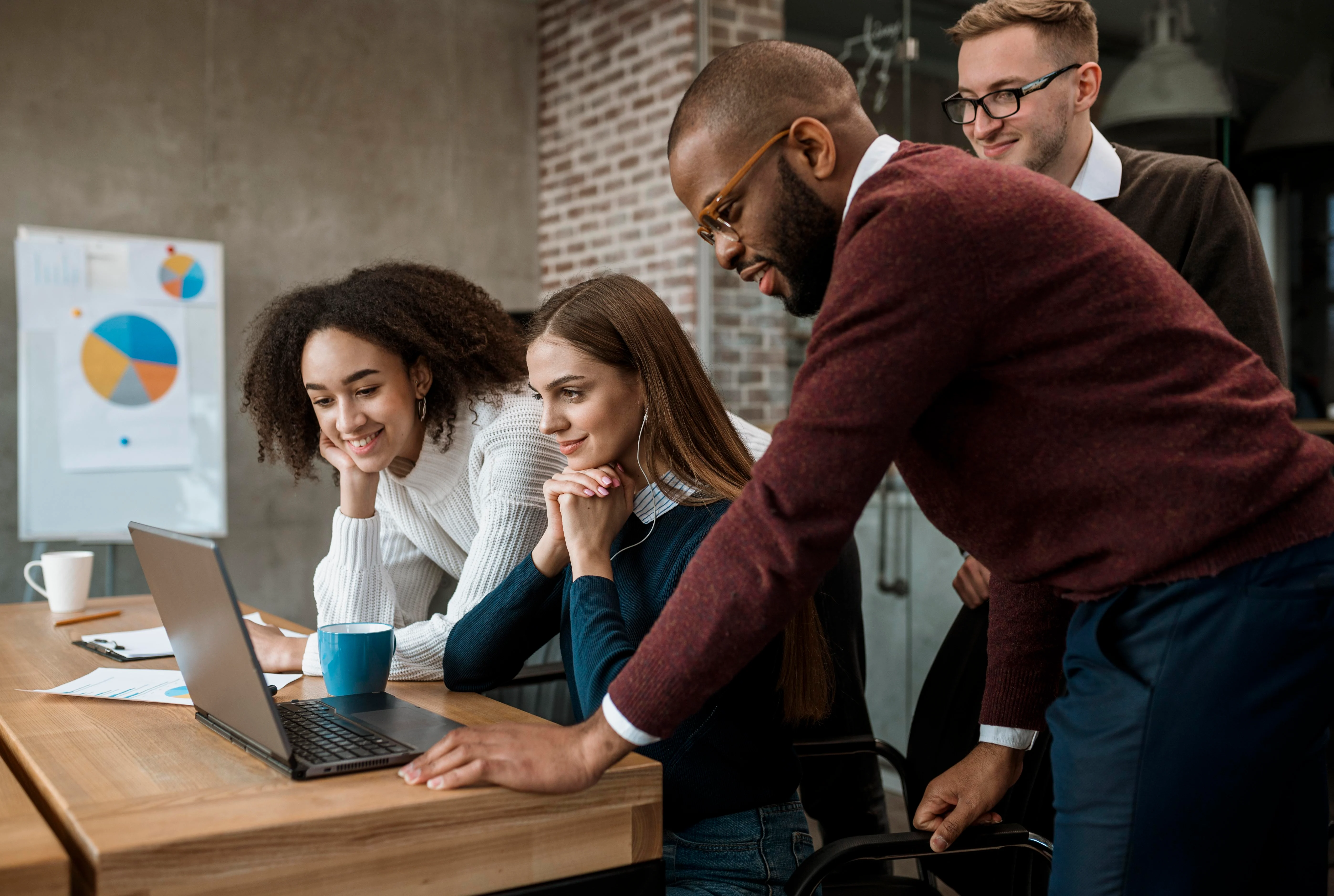Team Collaboration in a Modern Workspace A diverse group of four young professionals engaged around a laptop at a wooden table. Two women and two men are looking intently at the screen, displaying expressions of curiosity and focus.