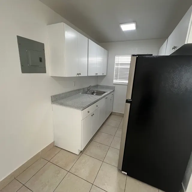 A compact kitchen featuring white cabinets, a stainless steel sink, and a black refrigerator, with tiled flooring and natural light.