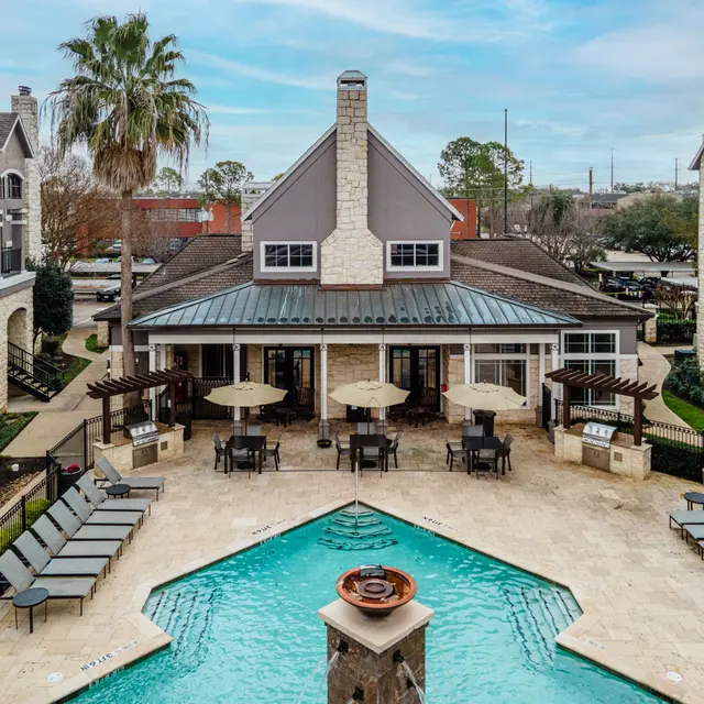Aerial view of a modern apartment complex with a central pool area. The pool is surrounded by lounge chairs and features a fountain at the center. There are shaded seating areas and palm trees in the vicinity.
