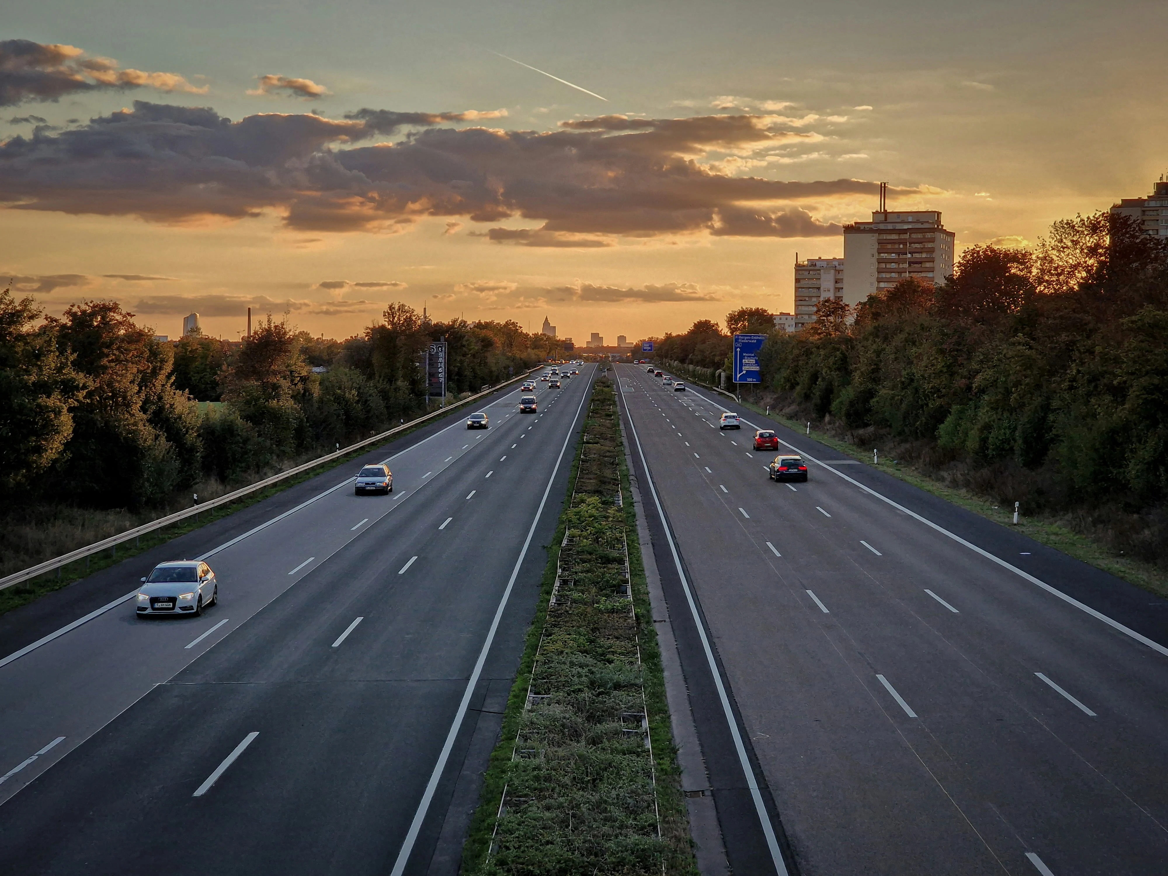 A long highway with multiple lanes at sunset, featuring several cars and greenery on the sides.