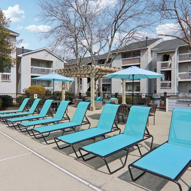 A modern outdoor swimming pool area with several light blue lounge chairs and umbrellas. In the background, there are residential buildings and greenery.