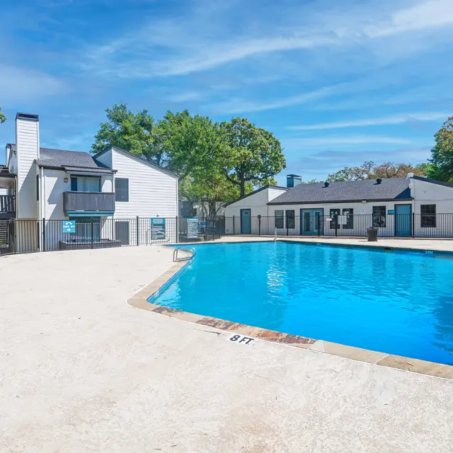 A clear blue swimming pool surrounded by a fenced area with apartment buildings in the background and sunny skies.