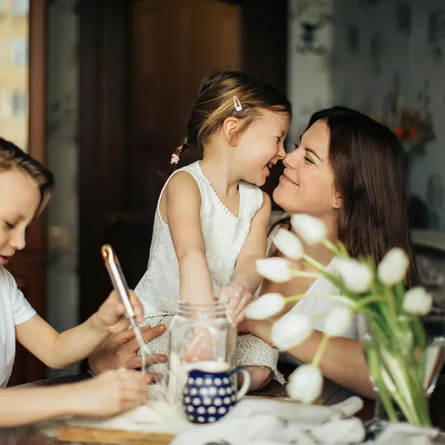 A mother interacts joyfully with her daughter while her son is engaged in an activity at the table.