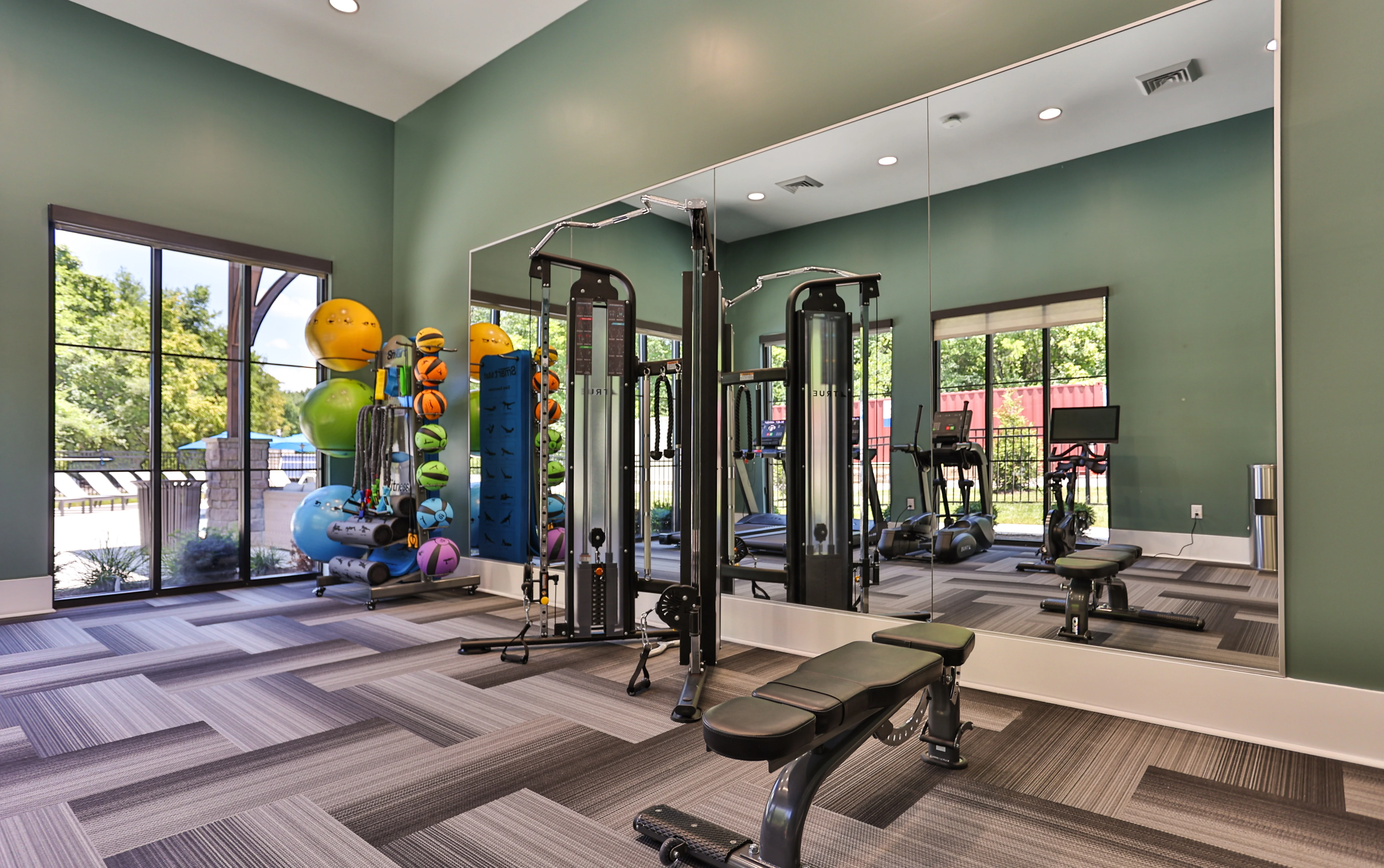 Modern gym interior featuring fitness equipment, weight bench, and mirrored wall with large windows overlooking green landscape.