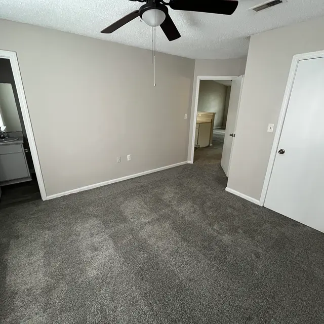 An empty room with light grey carpet, a ceiling fan, and neutral-colored walls. Two doors are visible, one leading to a bathroom area and the other to another room. Bright lighting from a window enhances the spacious feel of the room.