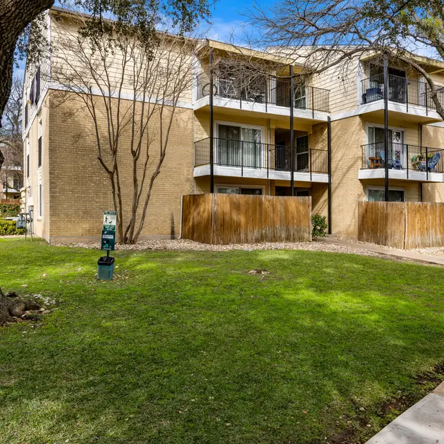 A multi-story apartment building surrounded by green grass and trees, featuring balconies and a wooden fence.