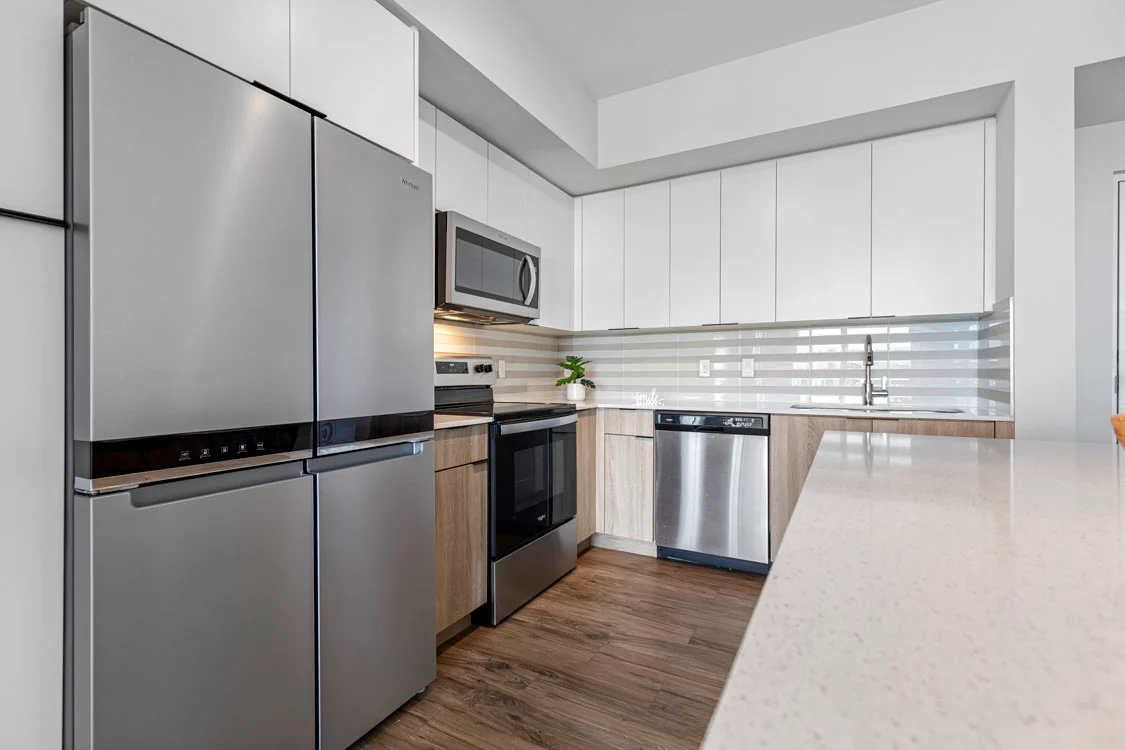 A modern kitchen featuring stainless steel appliances, including a refrigerator, microwave, oven, and dishwasher. The cabinetry is white with wooden accents, and the countertop is a light color. The backsplash is made of horizontal glass tiles, and there are potted plants on the countertop.