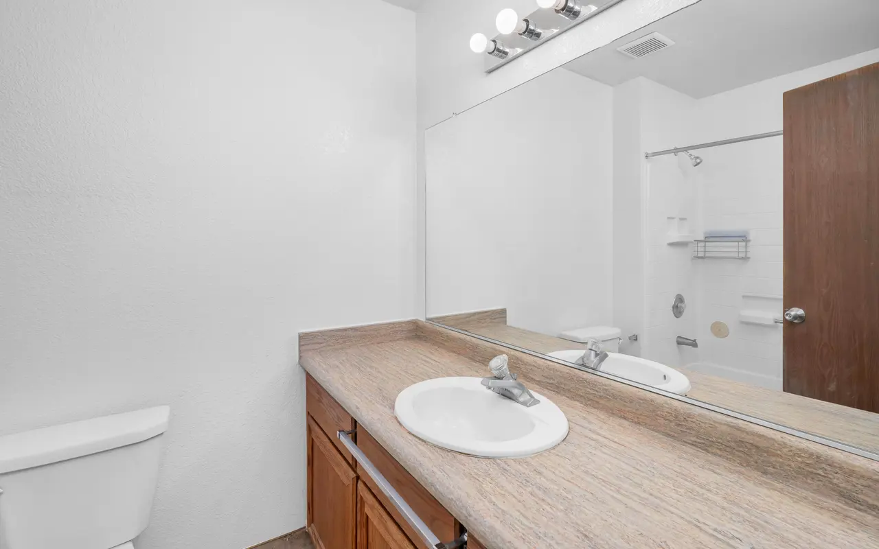 A clean and modern bathroom featuring a wooden vanity with a sink and mirror above, a white toilet on the left, and a shower area visible in the background.