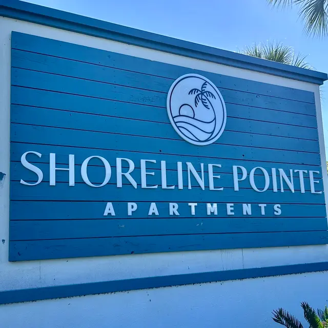 A sign for Shoreline Pointe Apartments featuring a stylized logo of a palm tree and waves, set against a blue wooden background.