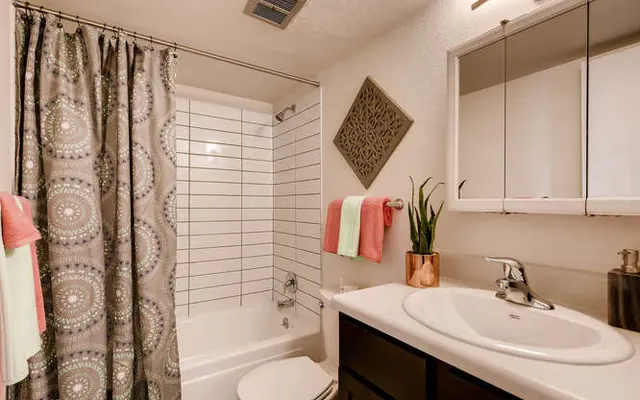 A modern bathroom featuring a white bathtub with shower, tiled walls, a shower curtain with a decorative pattern, and a sink with dark cabinetry. There are decorative towels and a small plant on the countertop.