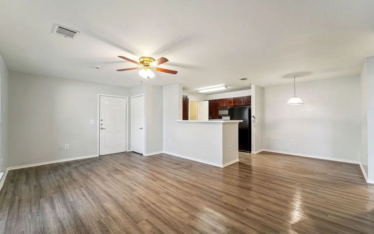 A spacious living area in a modern apartment with wooden flooring, a ceiling fan, and a well-defined kitchen space. The walls are painted light, and there’s ample natural light coming in.
