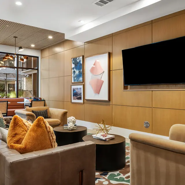 A stylish modern living room featuring a brown leather sofa and cream colored chairs with orange pillows, a round coffee table, and a wall-mounted television. There are decorative art pieces and a green lamp, with large windows revealing an outdoor area.