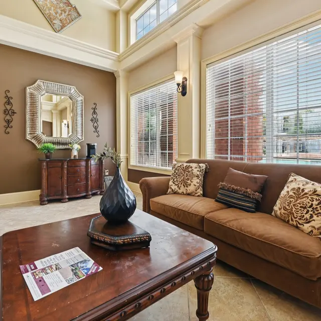 A stylish living room featuring a brown couch with decorative pillows, a wooden coffee table, and a large mirror on the wall. Natural light floods in through large windows, creating a welcoming atmosphere.