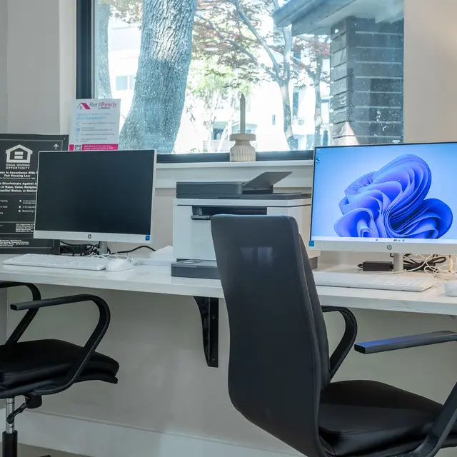 A clean office workspace featuring two black office chairs, two monitors on a white desk, a printer, and a well-lit window with trees outside.