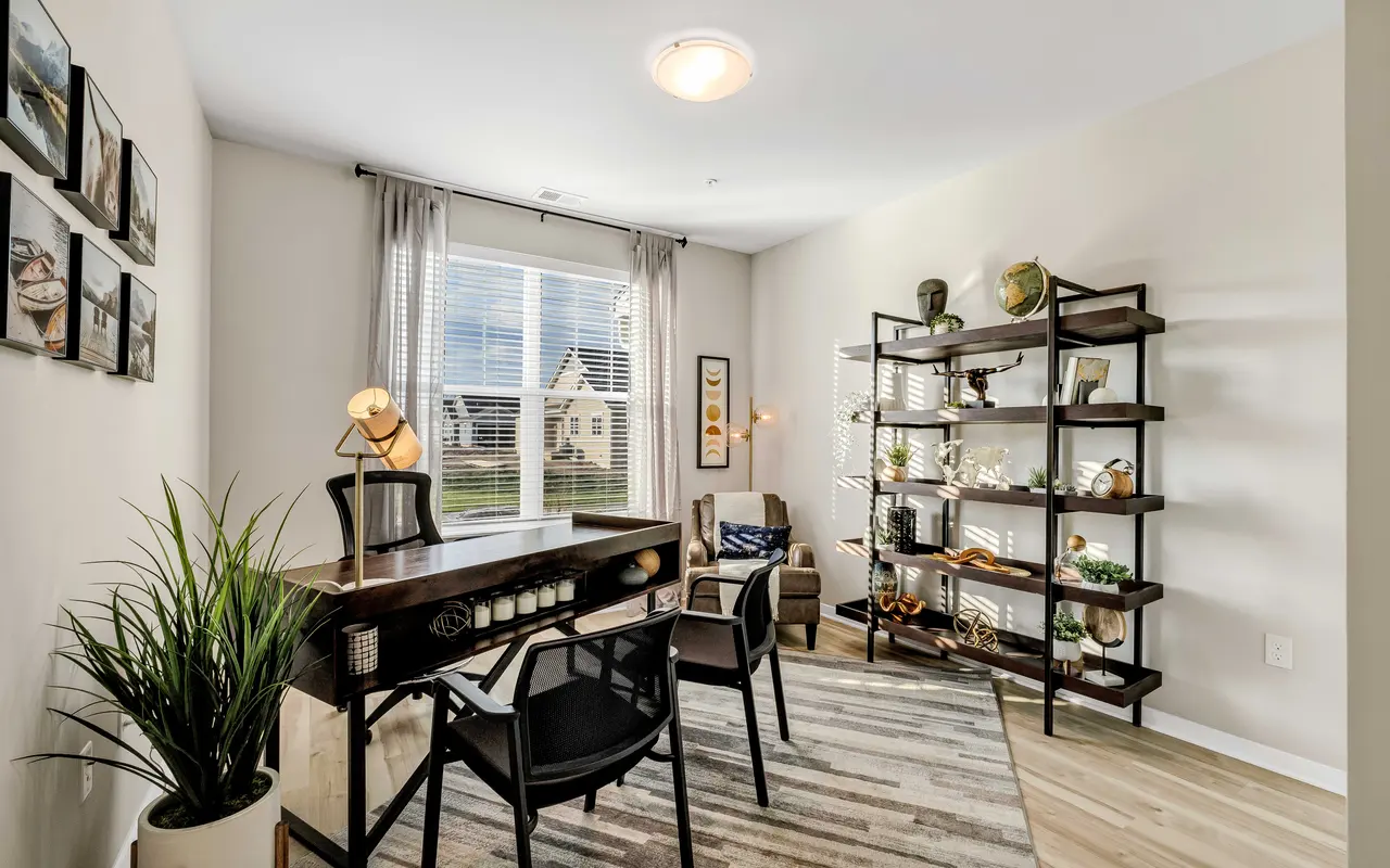 A modern home office featuring a wooden desk, black chairs, a tall shelving unit decorated with various items, and large windows with curtains allowing natural light to enter.