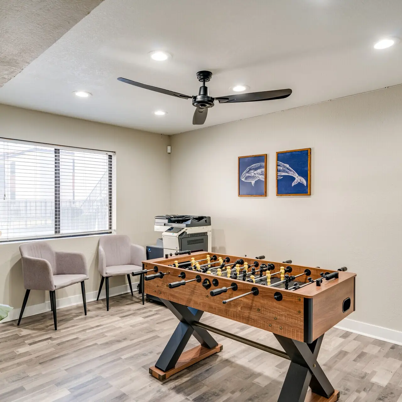 A modern game room featuring a foosball table, two chairs, a ceiling fan, and framed artwork of dolphins.