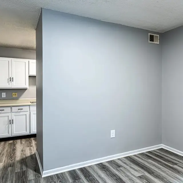 An empty room with gray walls and gray laminate flooring, featuring a small kitchenette area with white cabinets in the background.
