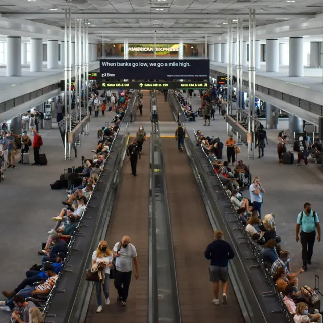 A wide view of an airport terminal with travelers walking and sitting. There are moving walkways and flight information screens above.