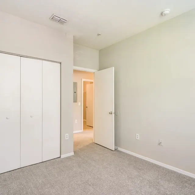 A spacious, empty room with light gray walls and carpeting. There are double doors leading to a closet on the left and an open door to another area on the right.