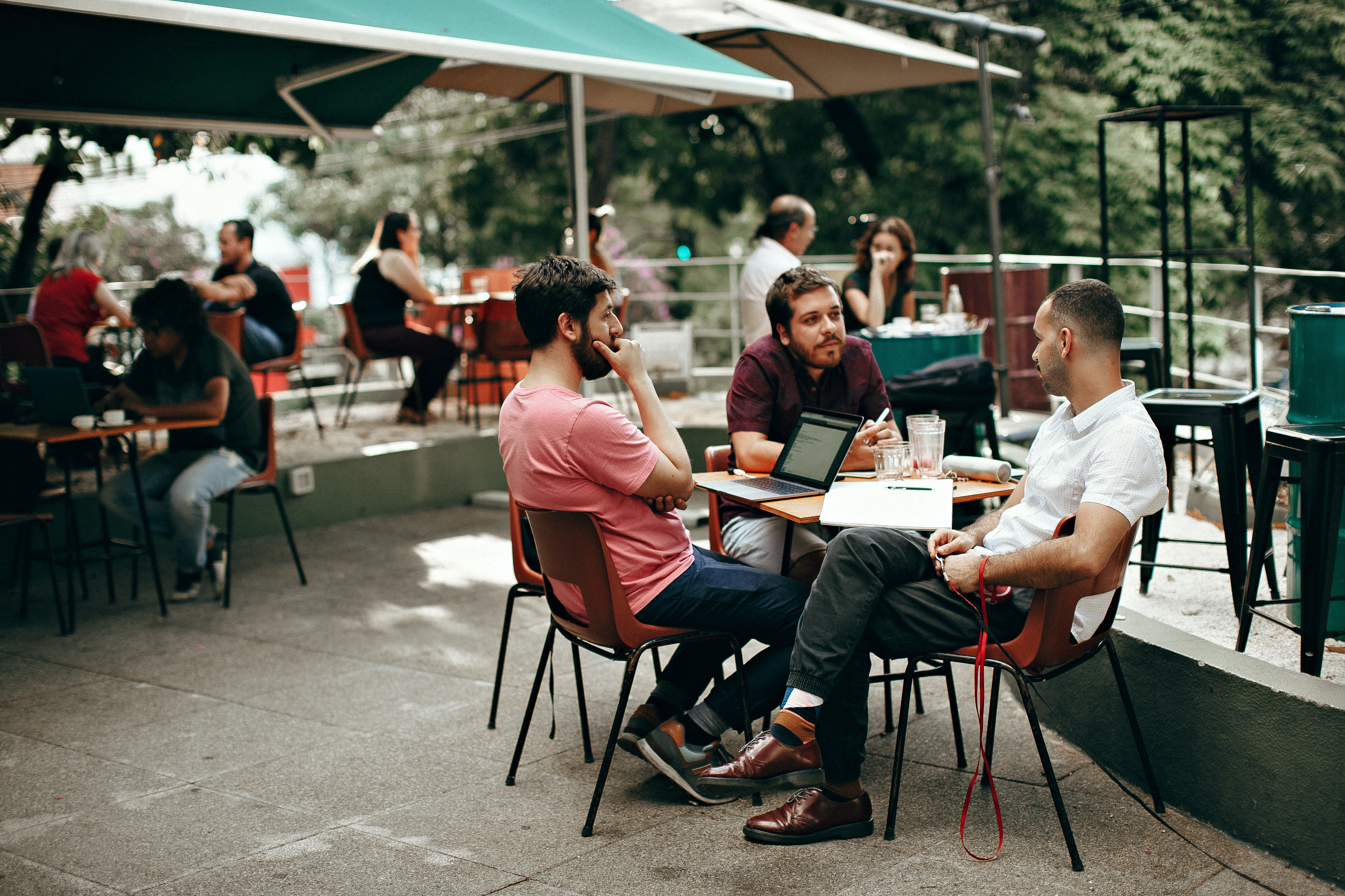 A group of three men sitting at a table outdoors in a cafe, engaged in conversation. One man is using a laptop, while the others are looking on. In the background, several other patrons are visible enjoying their time. There's greenery and shaded seating area.