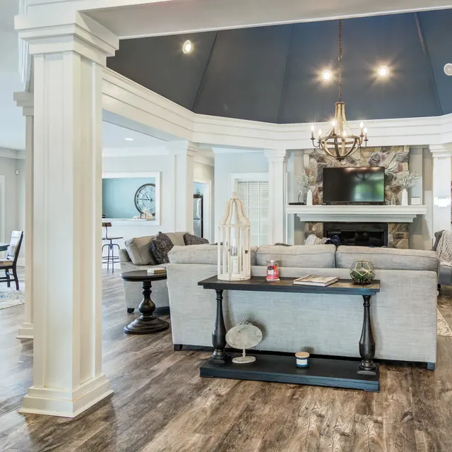 A spacious and modern living area featuring a gray sofa and a low table, surrounded by white columns and a dark ceiling. In the background, a stylish dining area is visible, along with a television above a fireplace.