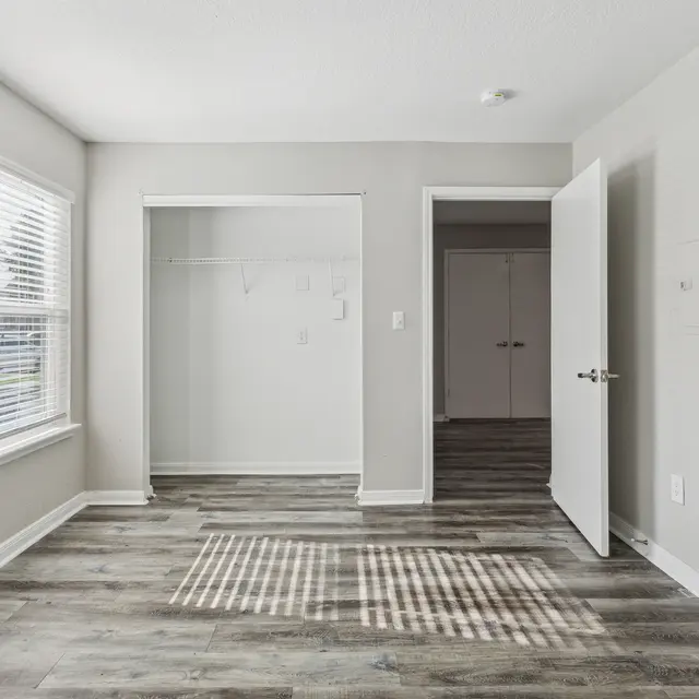 Empty Room Interior A light-colored, empty room featuring gray walls, wooden flooring, and two windows allowing natural light. There is a closet space on the left and an open door leading to another room on the right.
