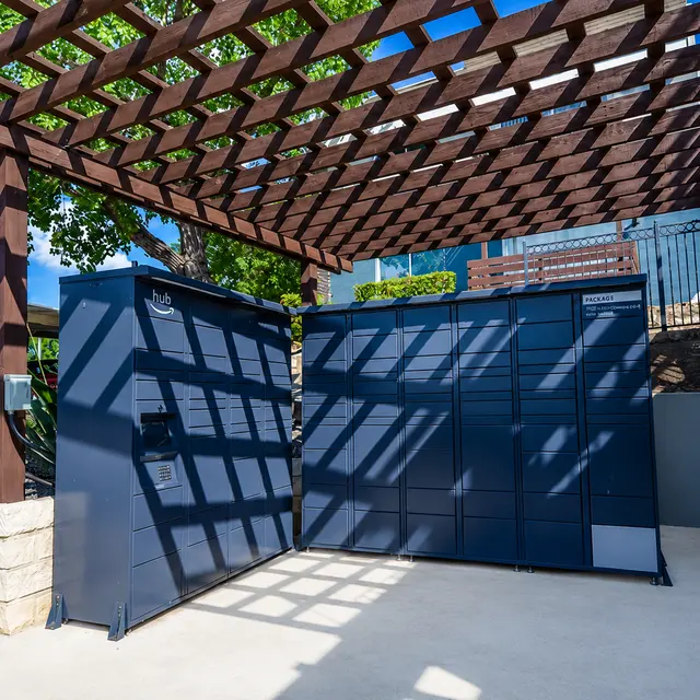 A modern package locker system located under a wooden pergola in a sunny outdoor setting, surrounded by greenery.