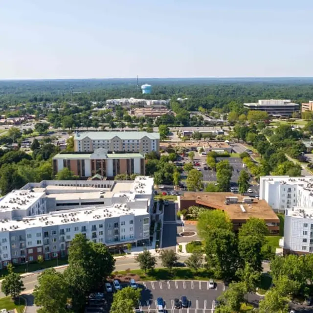 Aerial view of a cityscape featuring several buildings, parking lots, and green trees, with a clear sky above.