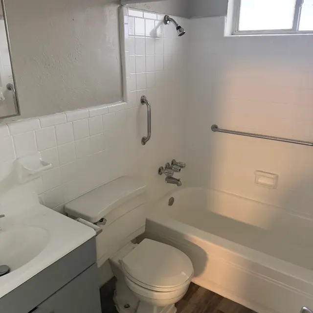 A small bathroom with a shower and bathtub combination, a toilet, and a sink with cabinetry. Natural light enters through a small window.