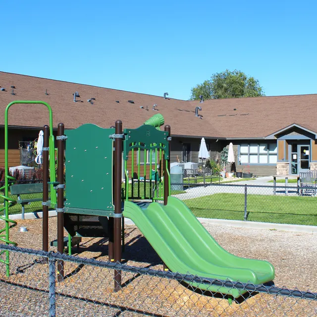 A colorful playground featuring green slides and climbing structures, surrounded by a fenced area with a clear blue sky in the background.