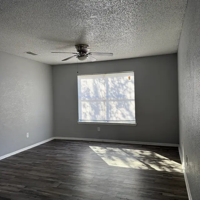 A spacious empty room with a ceiling fan, light gray walls, and dark wooden flooring. A window allows sunlight to filter in, casting shadows on the floor.