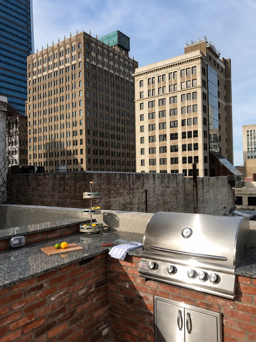 Rooftop Grill Area in the City A rooftop grill area featuring a stainless steel barbecue grill and a small wooden table with ingredients, overlooking tall buildings under a clear blue sky.