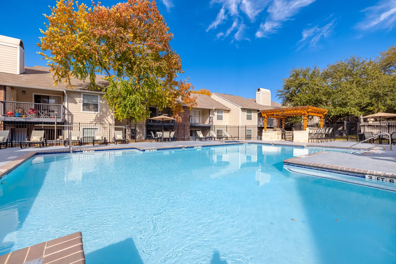 A clear blue swimming pool surrounded by residential buildings and a gazebo, with autumn foliage nearby.