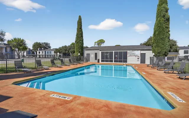 A clear swimming pool surrounded by lounge chairs, with a fenced area and a building in the background.