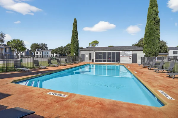 A clear swimming pool surrounded by lounge chairs, with a fenced area and a building in the background.