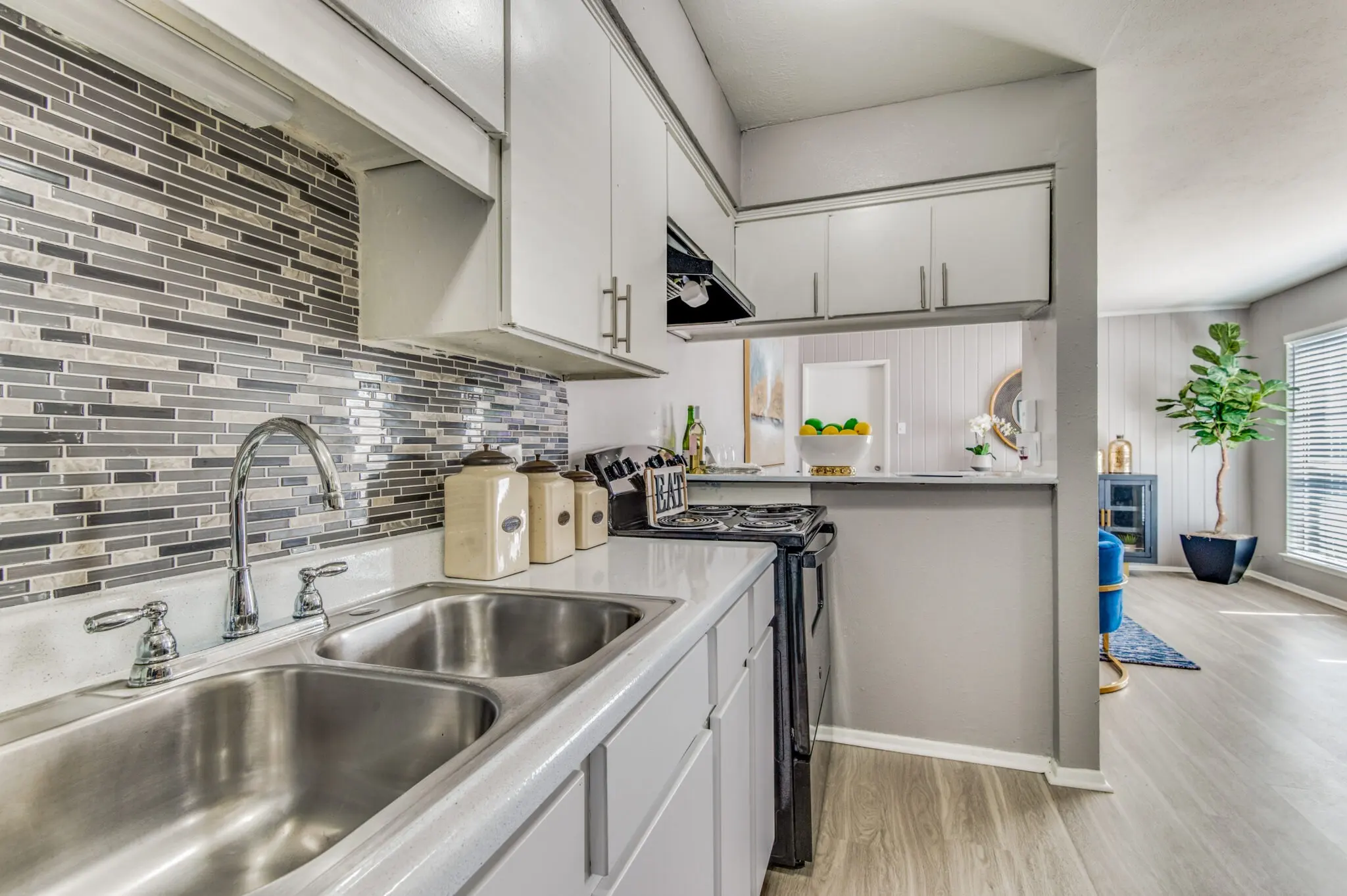 A spacious kitchen featuring dual sinks, a modern backsplash with gray tiles, and white cabinetry. The kitchen opens up to a living area with natural light and potted plants.