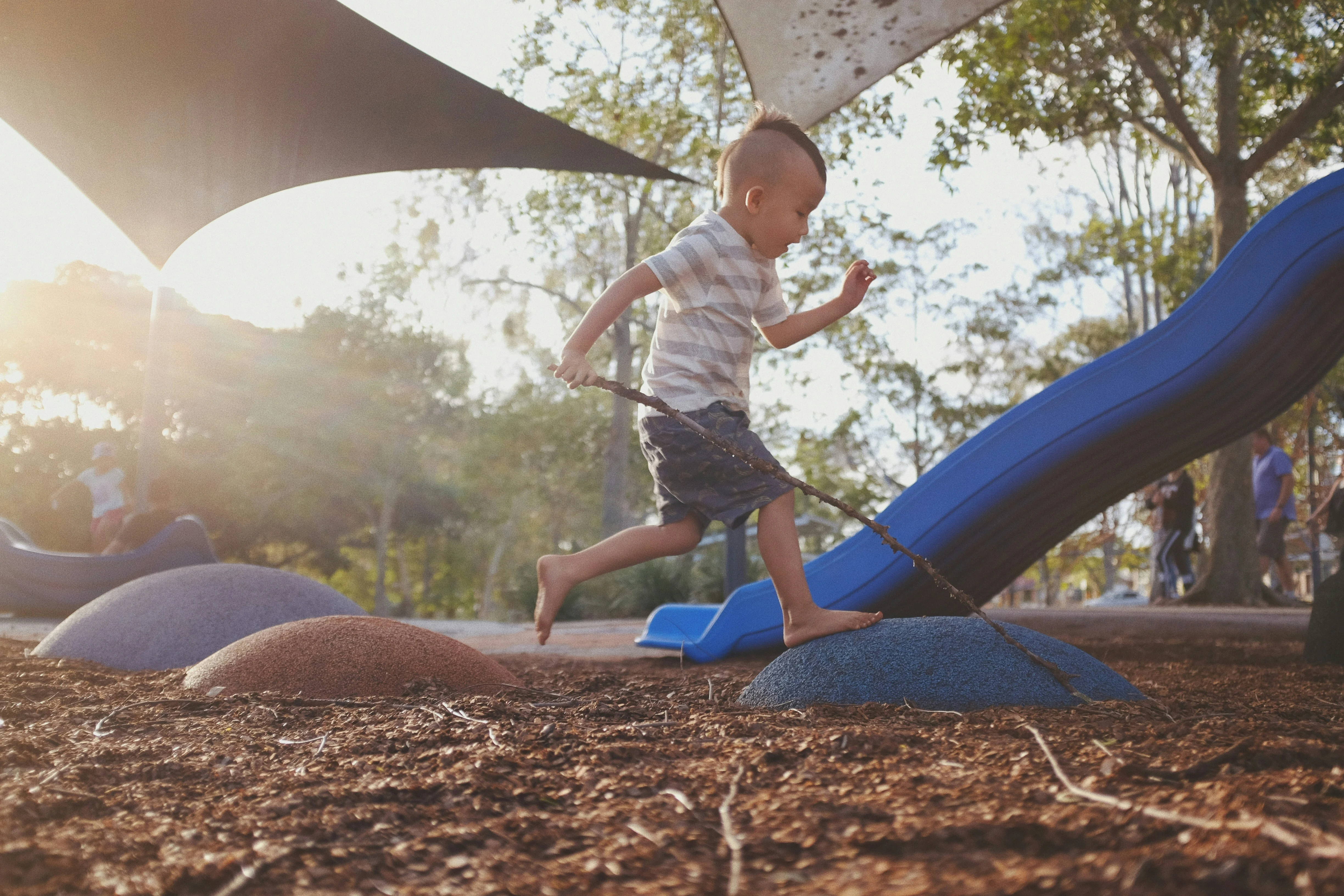 Child Playing at the Playground A young child running barefoot on a playground, holding a stick, with a slide in the background. The scene is filled with natural light and greenery.