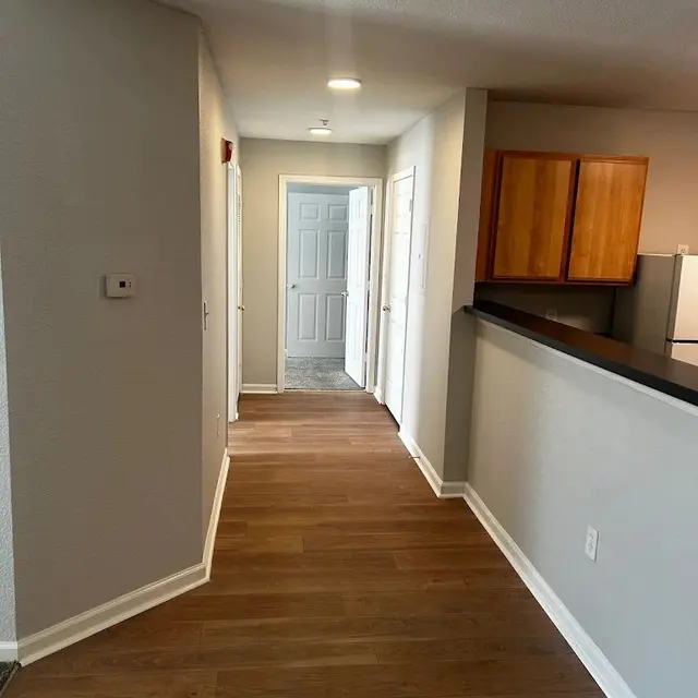 A modern apartment hallway featuring light wooden flooring, painted walls, and a kitchen area visible on the right with wooden cabinets. A white door is at the end of the hallway, and there are several doors on the left side, leading to different rooms.