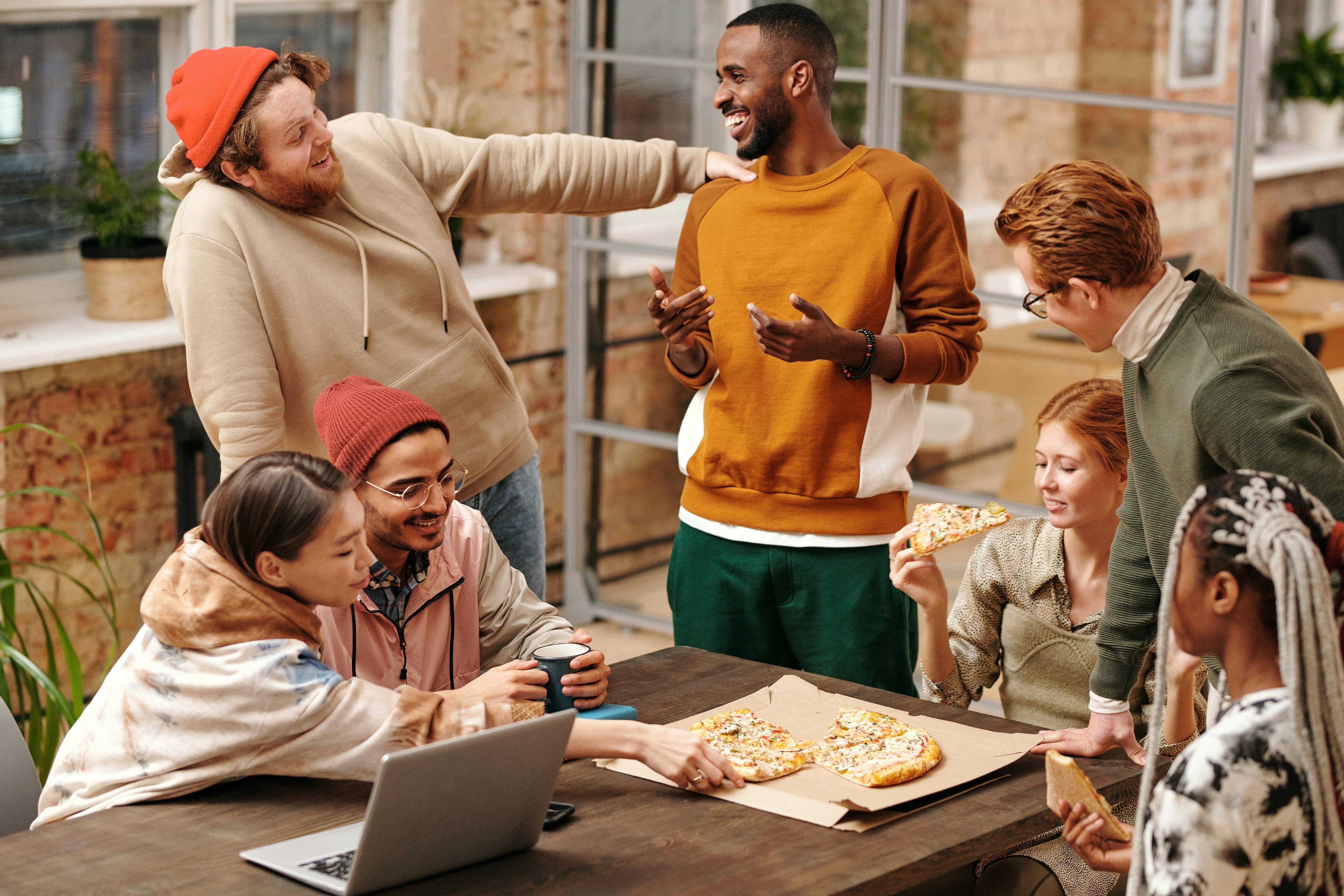 A diverse group of friends gathered around a table with pizza. They are engaged in lively conversation, with some holding slices of pizza and others drinking coffee. The setting is a cozy and modern workspace with large windows and plants in the background.
