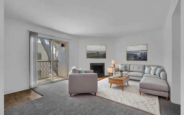 A modern living room featuring a light gray sectional sofa, a small coffee table, and a cozy armchair. The space is bright with natural light coming through sliding glass doors that lead to a balcony. Two framed paintings adorn the walls, and a light-colored rug covers the floor.