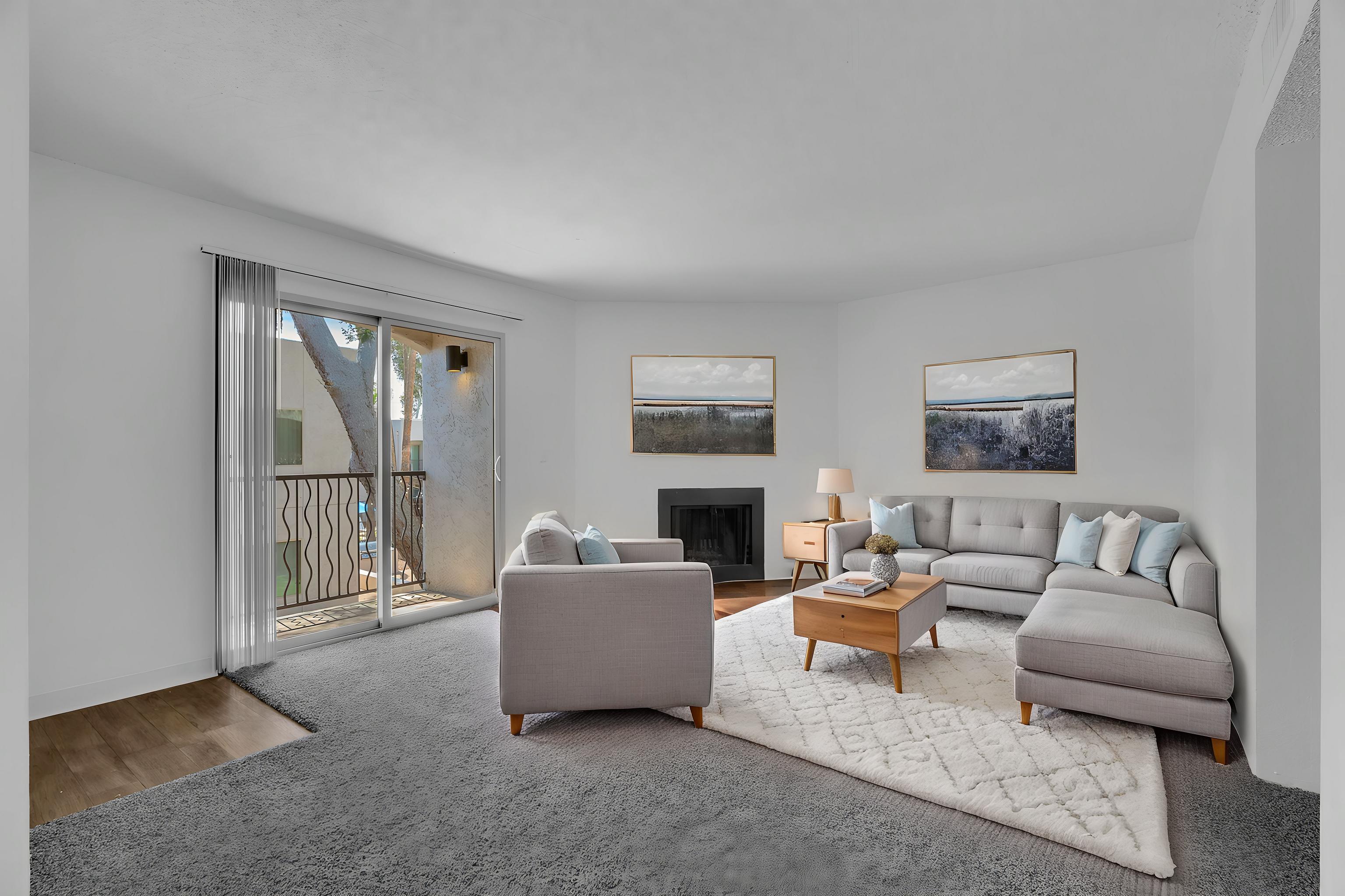 A modern living room featuring a light gray sectional sofa, a small coffee table, and a cozy armchair. The space is bright with natural light coming through sliding glass doors that lead to a balcony. Two framed paintings adorn the walls, and a light-colored rug covers the floor.