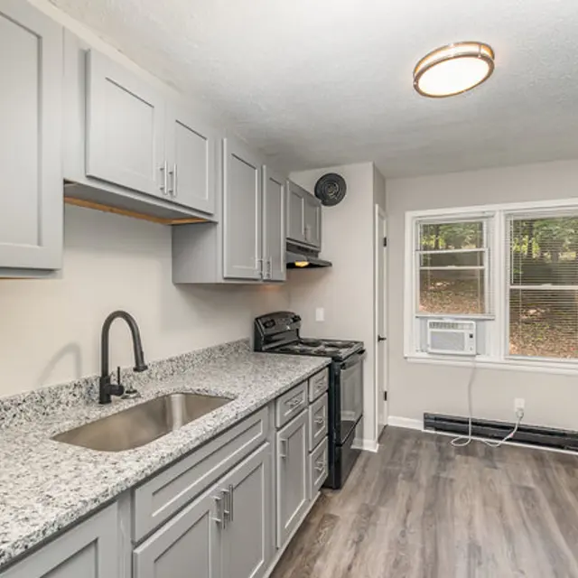 A modern kitchen featuring gray cabinets, a granite countertop, and stainless steel appliances including a stove and refrigerator. There is a window with natural light coming in, and an air conditioning unit is visible.