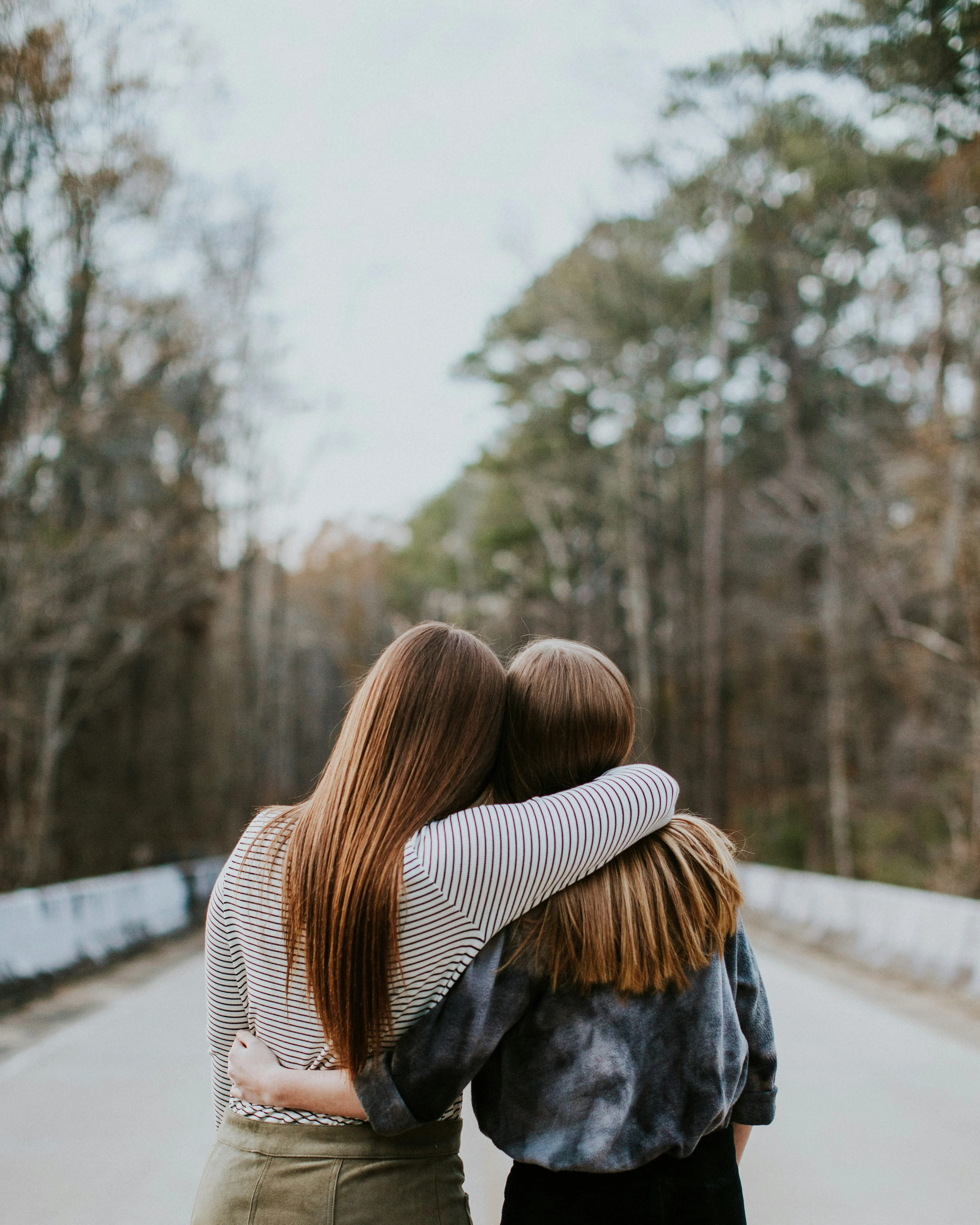 Friendship on a Quiet Road Two friends standing together, hugging each other from behind, facing a tree-lined road in a natural setting.