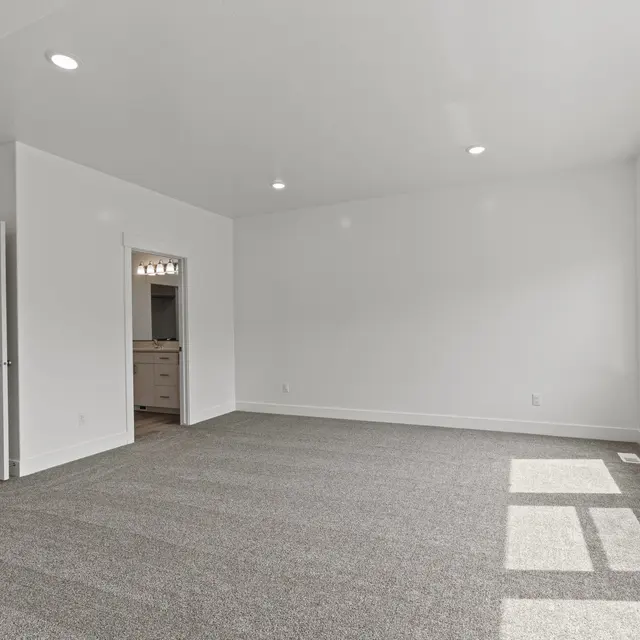 A spacious, empty living room with carpet flooring and white walls. It features large windows allowing natural light to enter, along with doors leading to another room.