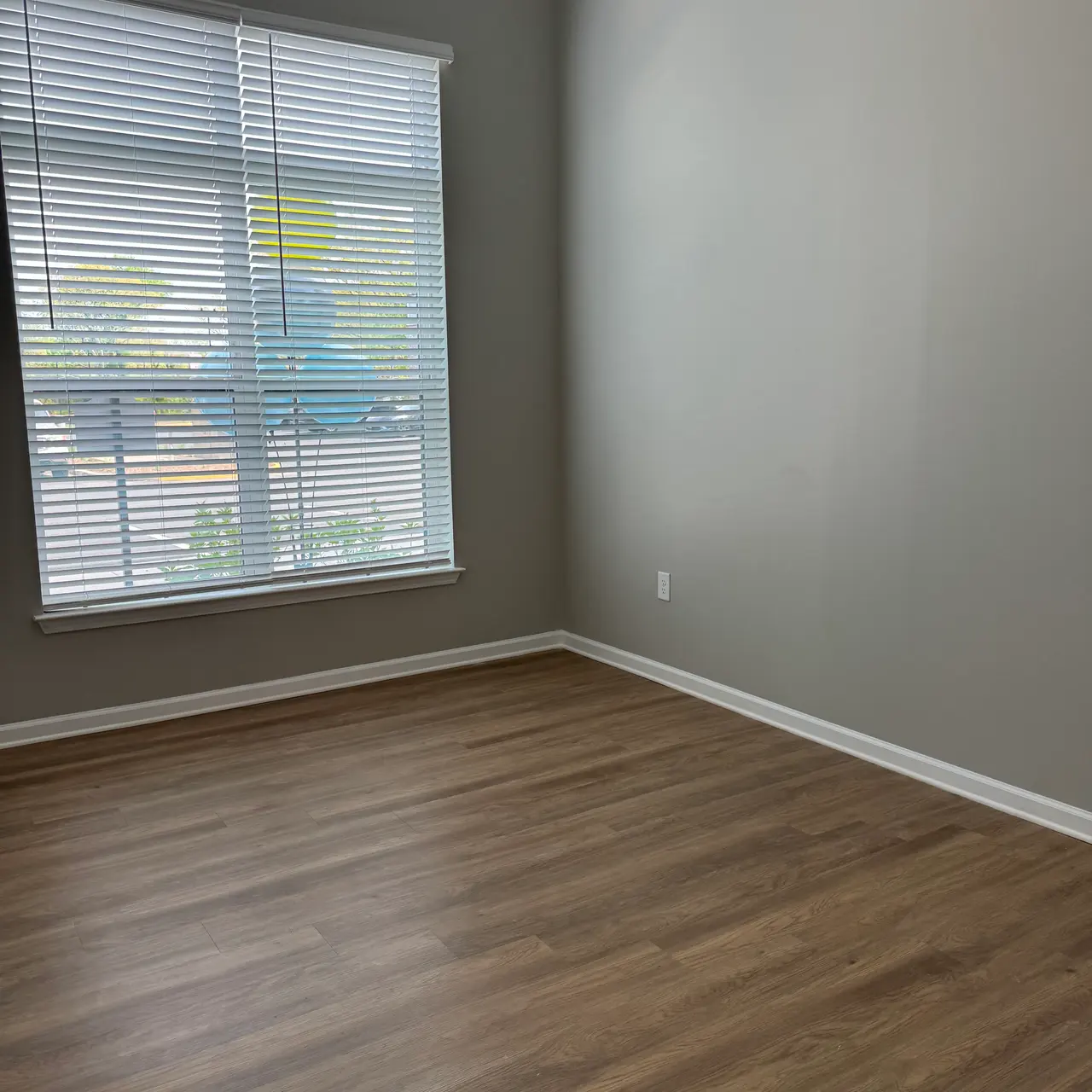An empty room with wood flooring and a window covered by blinds. The walls are painted in a light gray color, and the window allows natural light to enter.