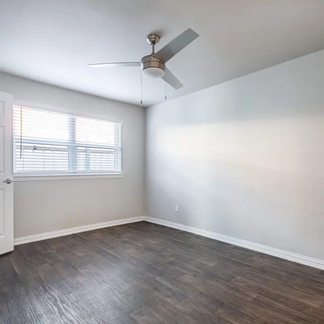 A spacious, empty room with a ceiling fan, hardwood-style flooring, and a window with blinds.