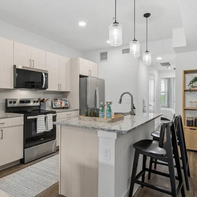 Contemporary Kitchen Interior A modern kitchen featuring light wooden cabinets, a stainless steel refrigerator, and a large countertop. Bar stools are arranged nearby, and the décor is minimalistic with a bright and airy feel.
